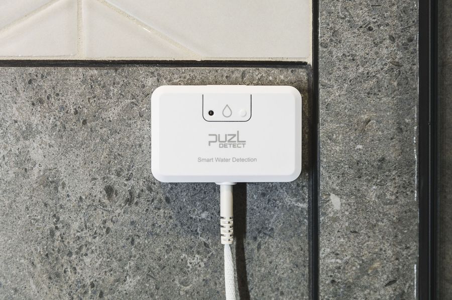 PuzL Detect mounted on a bathroom wall beside the shower, cable routed along the floor line